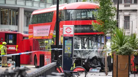 PA Media Emergency services at the scene following an accident involving a double-decker bus. The front of the bus' windscreen is smashed and the words on the bus read 'Hampstead Heath'.