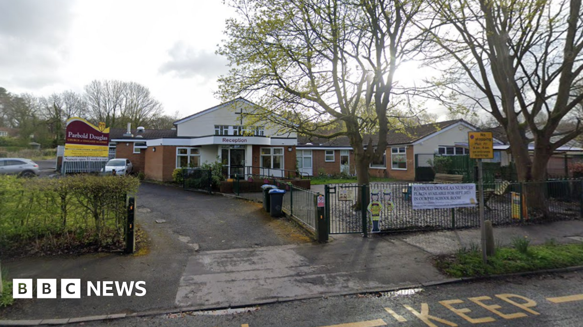 A Google Streetview of the exterior of Parbold Douglas Church of England Academy. It is a single-storey building with a Christian cross on the outside. There is a sign outside the school in the car park.
