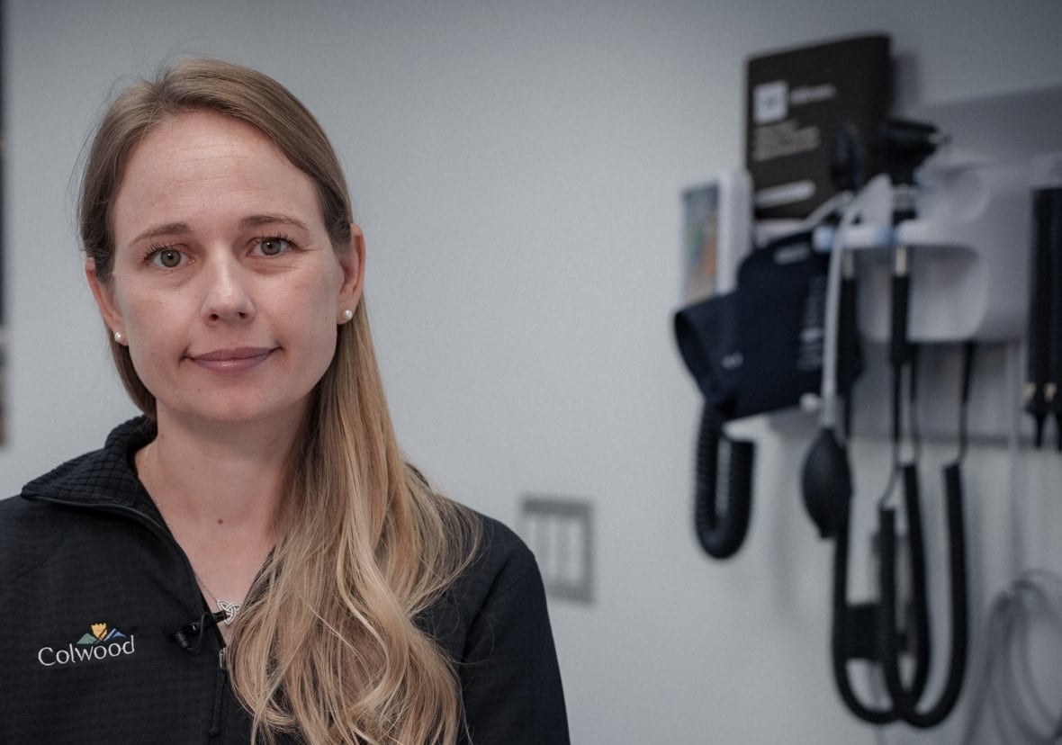 a woman with blonde hair smiles for the camera, as she sits in a doctrs office where stethoscopes and other medical tools are visible on a wall behind her