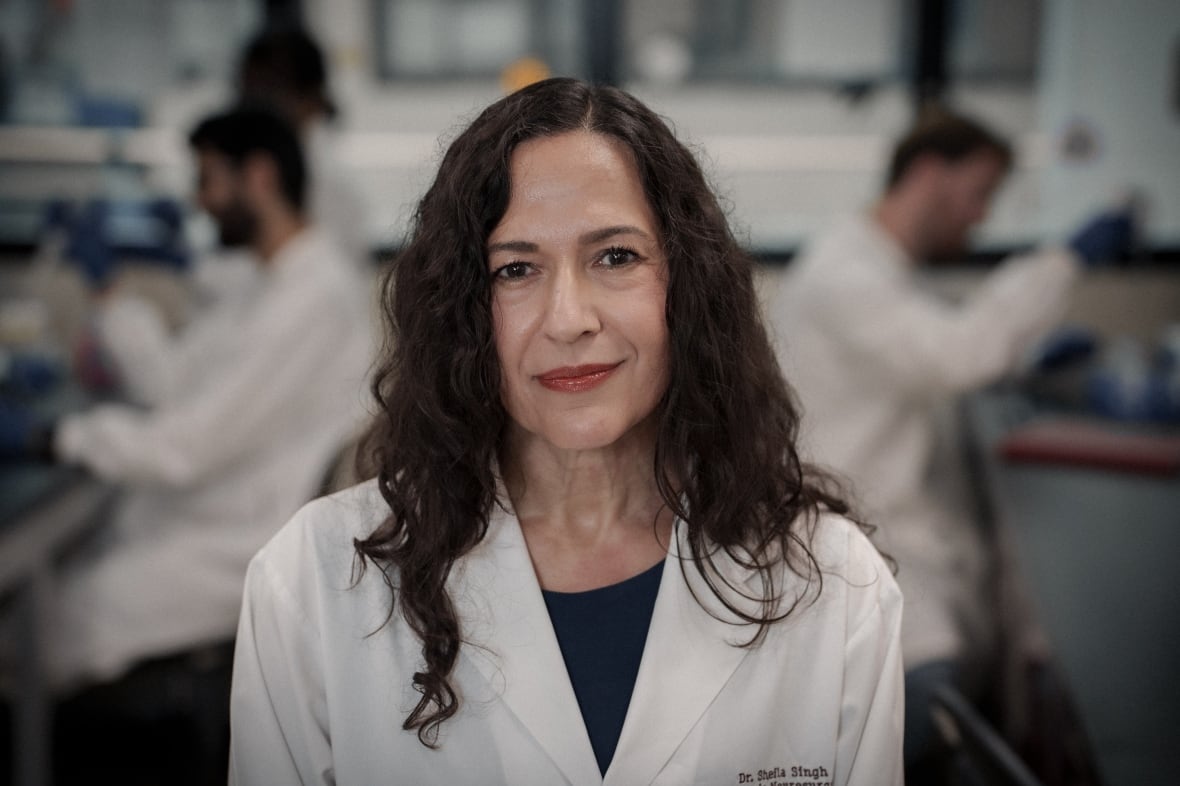 A woman with curly, brown hair past her shoulders wearing an embroidered lab coat stands with two scientists working at lab benches on both sides of her in the background.