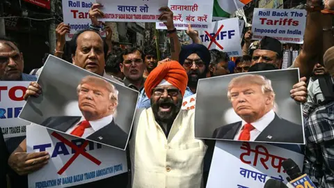 AFP via Getty Images A man wearing an orange turban and white top holds up two pictures of Donald Trump and shouts angrily at the camera. Behind him are a crowd of men holding up signs stating "roll back the tariffs imposed on India".