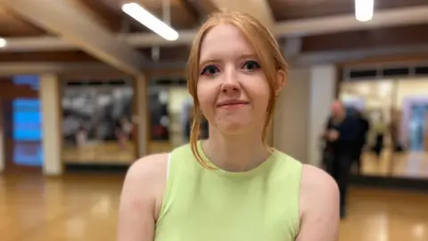 Amy has ginger hair which she is wearing pulled back, except for 2 strands which frame her face. She is wearing a tank top which is a pale shade of green. She is standing in a studio lined with mirrors. The background around her is blurred. 