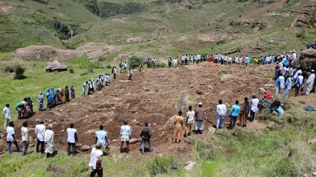 A group of people lining the edges of a large area of ground containing what appears to be freshly dug burial mounds