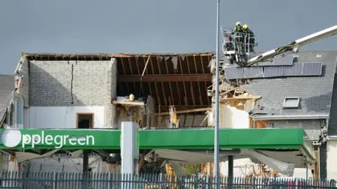 PA Media Two firefighters in an aerial platform appliance above the scene of the explosion at a service station in Creeslough in October 2022.  Most of the pitched roof of the front of the building has been blown off.  The inside of the wooden rafters are exposed, tiles are missing, solar panels have been displaced and there is a large crack in a grey brick wall which reaches the pitch of the roof. 