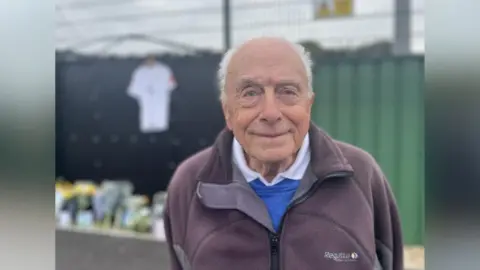 George Carden/BBC Michael Stemp pictured at the Chichester City FC football ground. Michael is wearing a white shirt, blue jumper and black zip up jacket. He has white hair. The floral and card tributes can be seen in the background, though are partially blurred out.