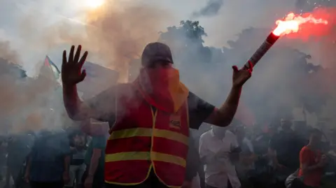 Getty Images A man in a hi-vis jacket and sunglasses waves a lit, red flare at a protest in France