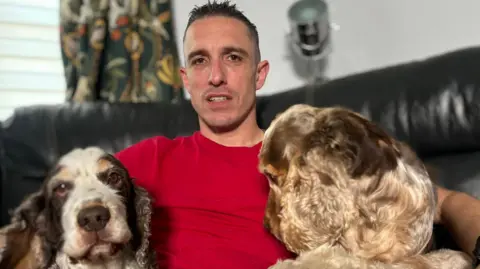 Matt Precey/BBC Dan Cooper in red T-shirt sitting on a black sofa in a living room. He has his arms around two dogs, one is looking directly at the camera. 