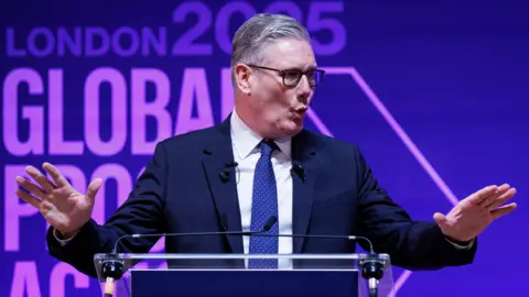 EPA A man with grey hair and glasses speaks at a glass podium, gesturing with two open hands. Behind him reads "London 2025: Global Progress Action Summit", emblazoned on a purple background.