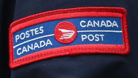 Bloomberg via Getty Images A close-up blue and red badge on a dark blue uniform sleeve with the Canada Post logo in both French and English. 