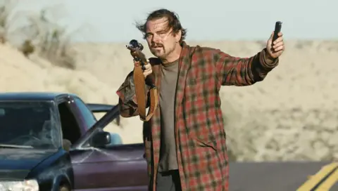 Warner Bros Leonardo DiCaprio as Bob in One Battle After Another standing outside a car holding a gun 