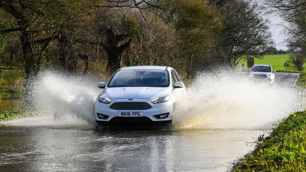 A photo of a car driving through flood water on a road
