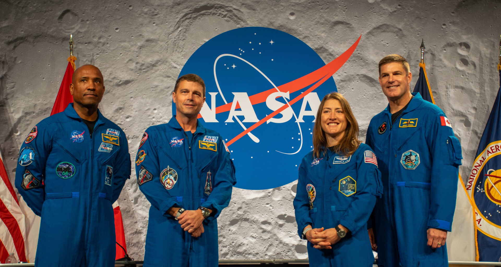 Four people wearing blue jumpsuits stand next to each other in front of a desk with a NASA logo behind them