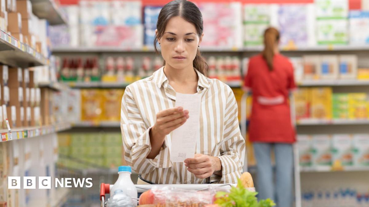 Young woman with dark hair in supermarket wearing beige and white striped shirt looking at receipt behind shopping trolley