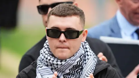 PA Media Liam Óg Ó hAnnaidh has short dark hair, he is wearing black sunglasses, a black and white scarf and a black coat. 
