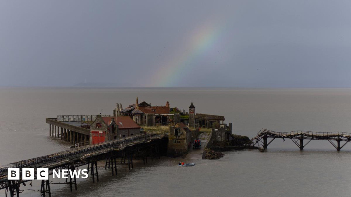 A pier that is falling apart is seen under a rainbow and grey sky.