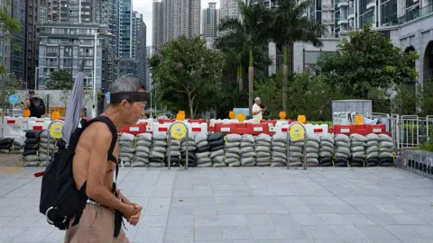 Getty Images A shirtless man wearing a headband and a backpack walks in front of a barricade made of sandbags in the street