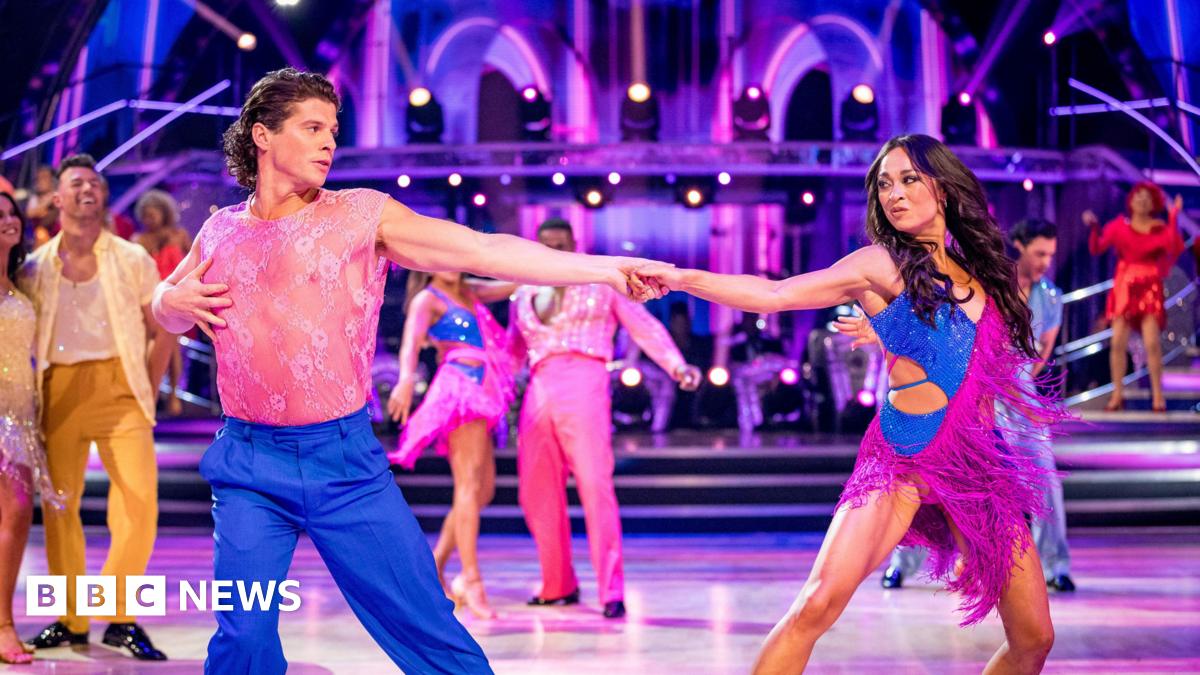 Lewis Cope & Katya Jones on the launch show of the new series of Strictly Come Dancing - the two are in the middle of the group dance, both wearing a dazzling combination of pink and blue, holding hand with their arms extended to maximum length, looking at each other in the middle of a dance move