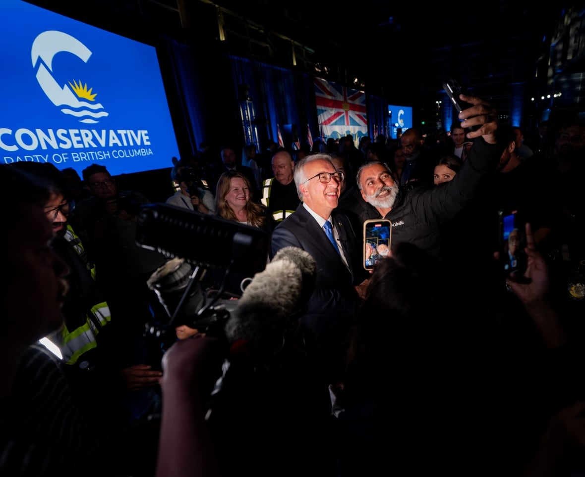 A person takes a picture with a man in front of a screen reading 'Conservative Party of British Columbia'.