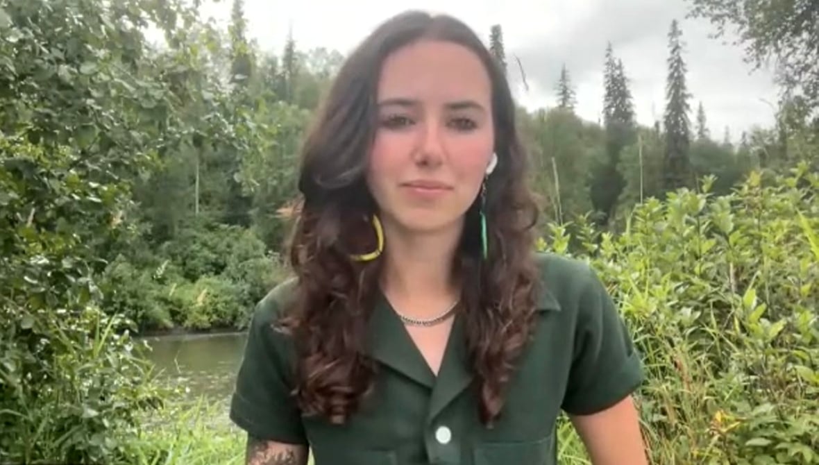 A woman in a green shirt stands outside in a forested area. 