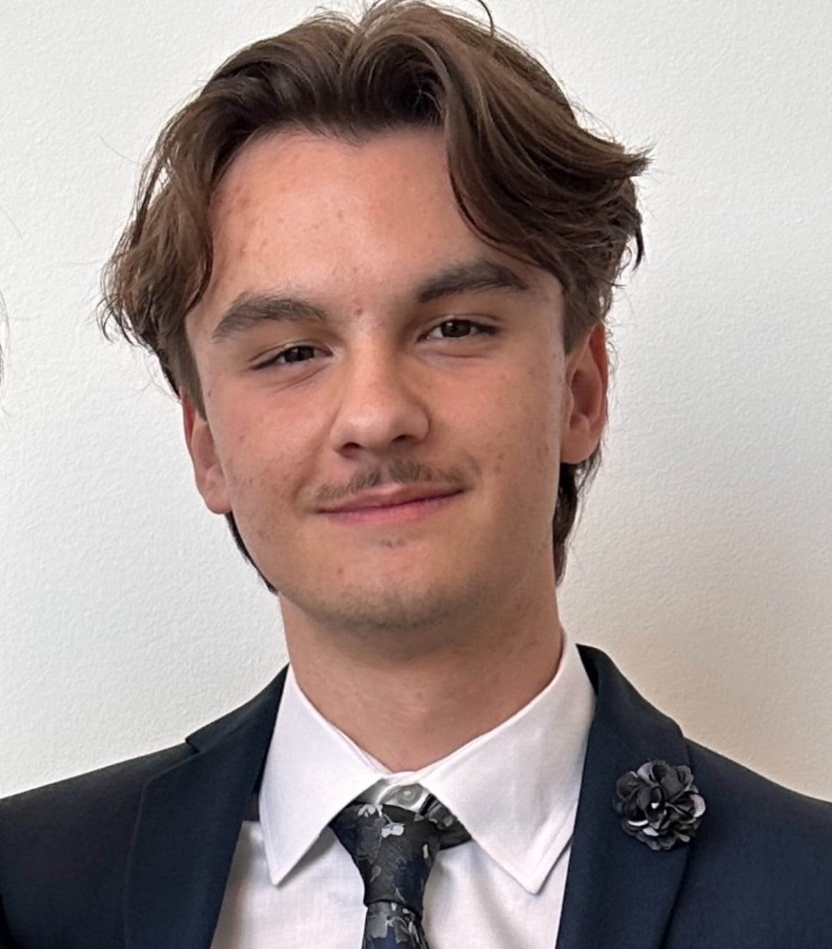 A young man with wavy brown hair wearing a blue suit and tie smiles at the camera