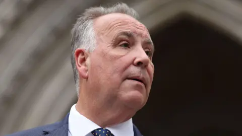 EPA/Shutterstock Chris Whitbread has short grey hair and is wearing a navy suit jacket, white shirt and navy tie with small yellow squares. He is standing outside a cream archway at the High Court.