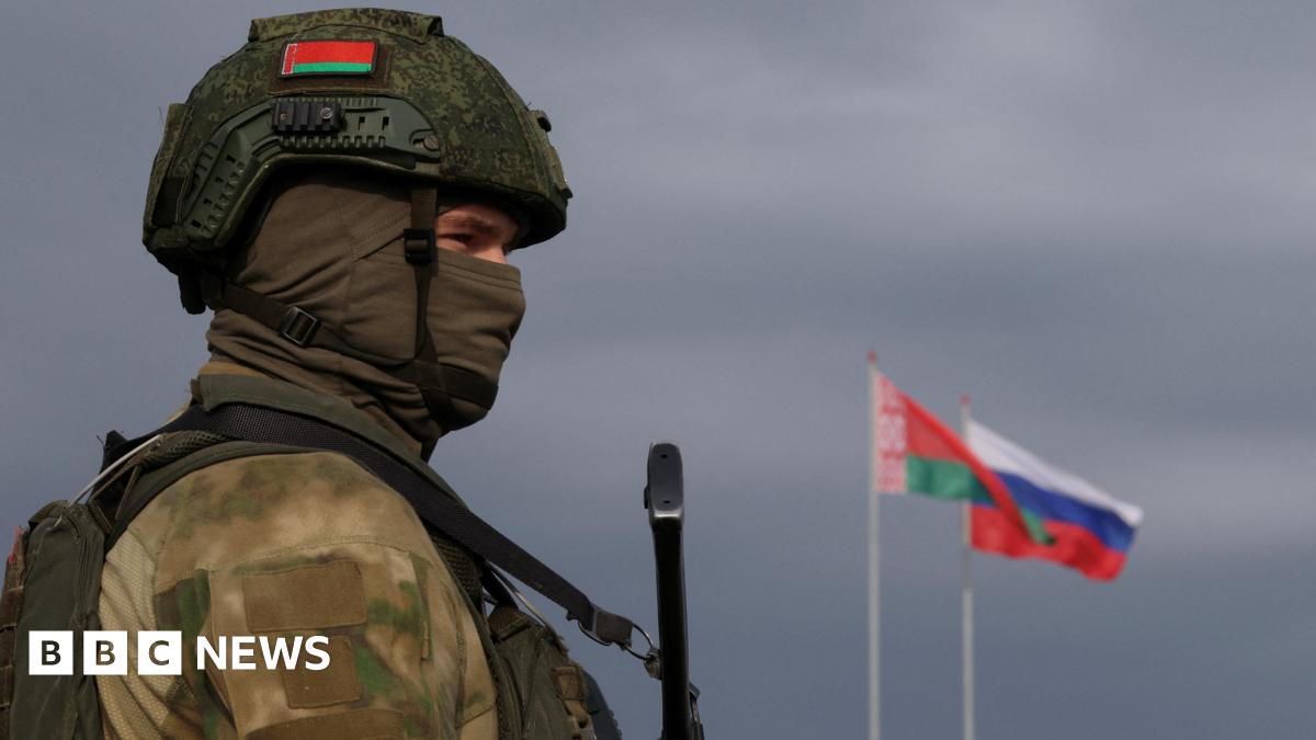 A Belarusian serviceman stands holding a rifle. There are a Belarusian and a Russian flag in the background