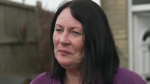 A woman with shoulder-length black hair wearing a purple top looks away from the camera. She appears to be standing outside her house.