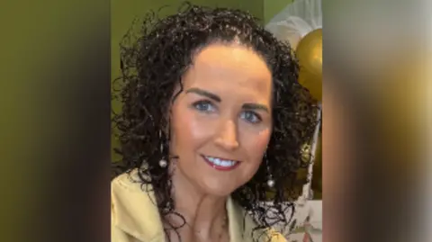 Family handout A woman with long black curly hair smiles at the camera as she wears a yellow blazer and gold necklace. She has blue eyes and is wearing make up. She is wearing gold dangly earrings with pearls on them.