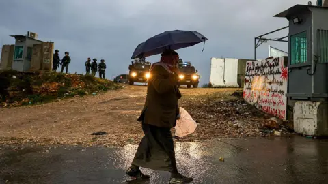 AFP via Getty Images A man walks through a checkpoint, holding an umbrella. 