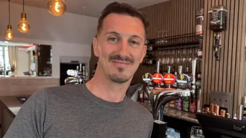 The Sporting King pub's owner Ian is propped up against the bar with a row of beer pumps over his shoulder  