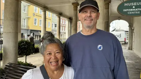 Smiling Californian couple Miriam and Taylor Prell are standing outside the Windsor Guildhall building just yards from the Castle. Smiling into the camera Taylor's wearing a blue baseball cap while Miriam has a shoulder bag slung over her white top.