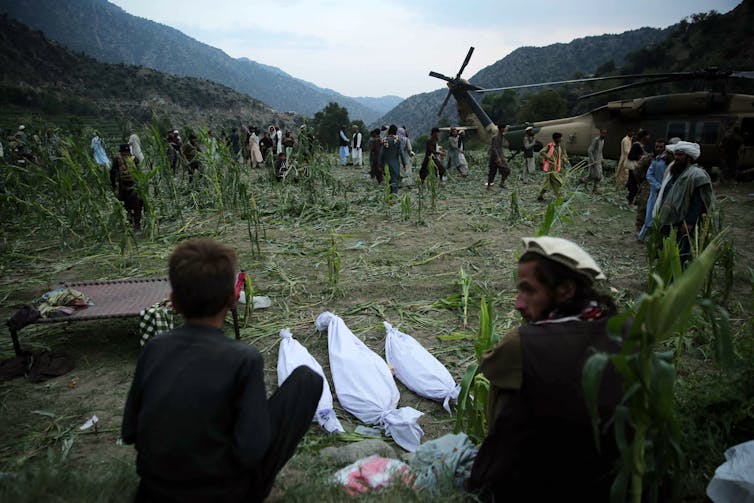 A helicopter in a field, next to a crowd of people and three bodies in white bags.
