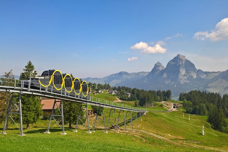 A yellow and black railway car travels along a track, with mountains in the background.