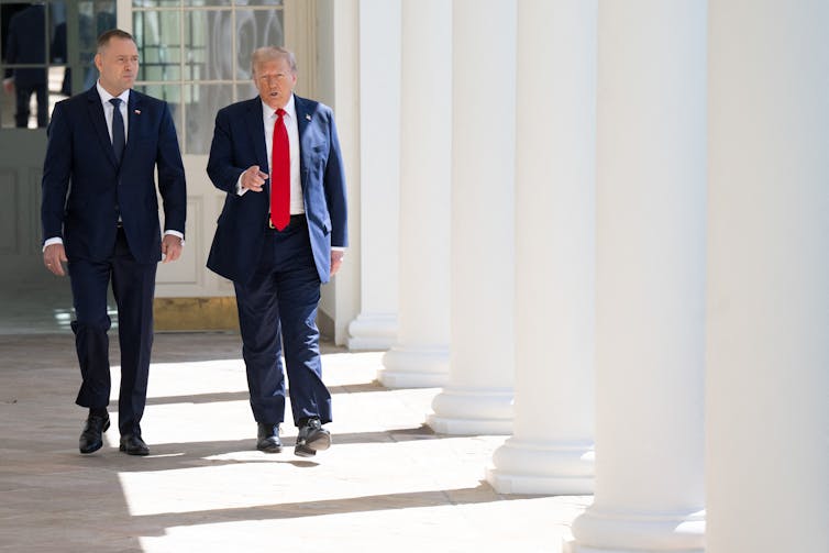 Two men in suits walk down a sunlit corridor.