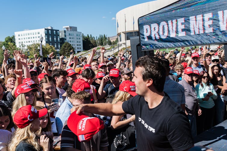 A young man wearing a black t-shirt extends his arm toward a crowd of young people, many of whom are wearing red hats.