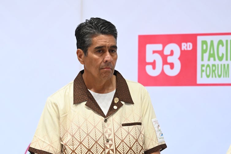 A man in a beige and brown short stands in front of a sign