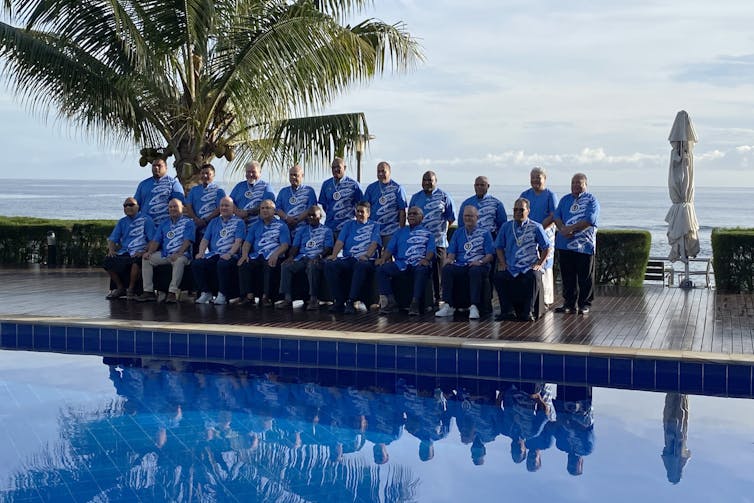 Two dozen people in blue shirts pose for a poolside photo.