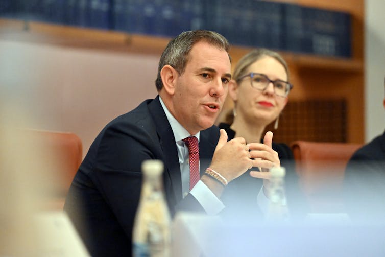 A man wearing a black suit and a red tie sitting at a table.