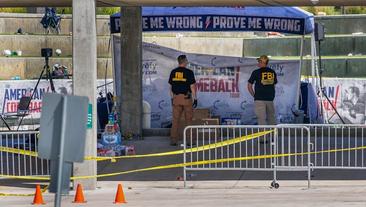 Two men wearing dark shirts with yellow writing stand behind a yellow roped off area that has signs that say 'American Comeback.'