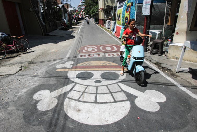 Two people on a moped drive over a mural showing a skull and crossbones in a hat.