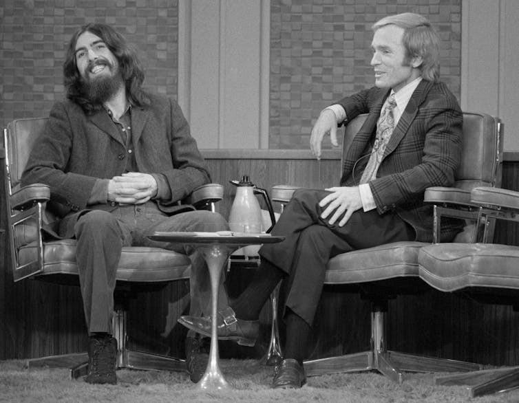 Black-and-white image of two men sitting in chairs and chatting on a television set.