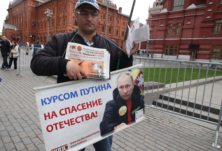 Man holding a sign with Vladimir Putin's face on it hands out newspapers.