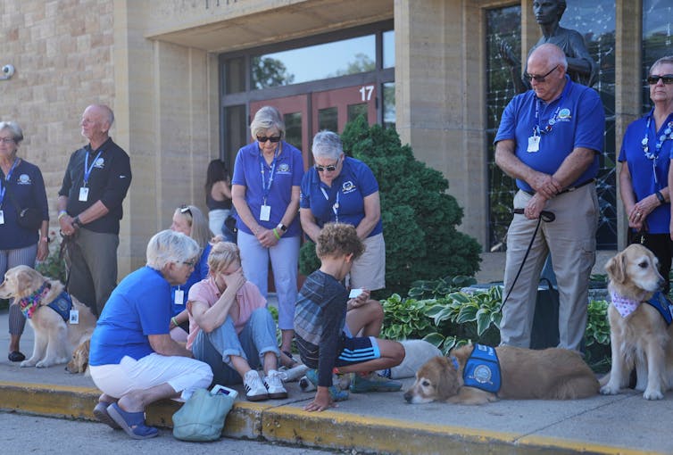 People in periwinkle blue T-shirts stand while children sit on the ground, surrounded by dogs.
