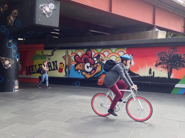 a cyclist and a walker emerge from beneath an overpass, which is painted with a mural