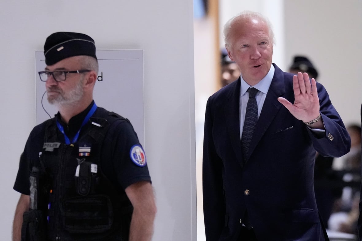 A balding white haired man wearing a suit and tie waves with left hand to people off camera as he walks in a hallway.