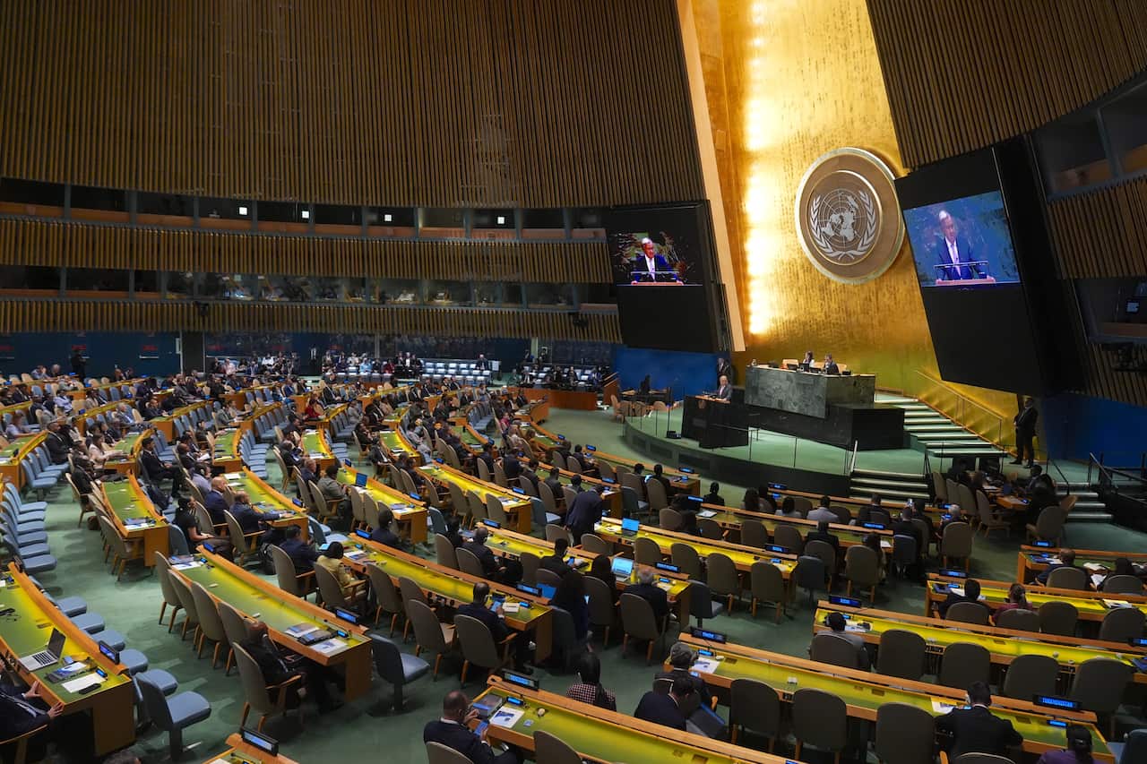 A wide-angle view of the United Nations General Assembly Hall shows a large group of people seated in curved rows, with an ornate, golden wall and two large video screens in the background. A speaker is visible at a podium on a raised stage, with his image displayed on the screens.