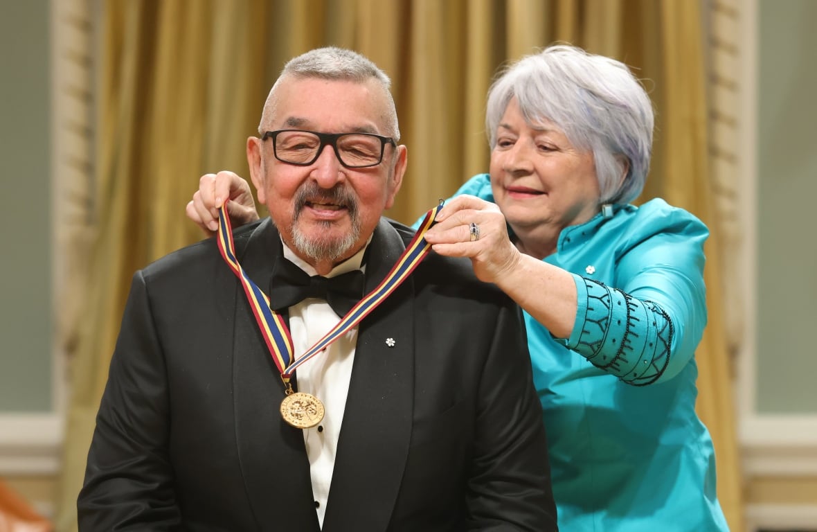 Man with grey hair and glasses has a medal placed around his neck by a woman wearing a blue dress