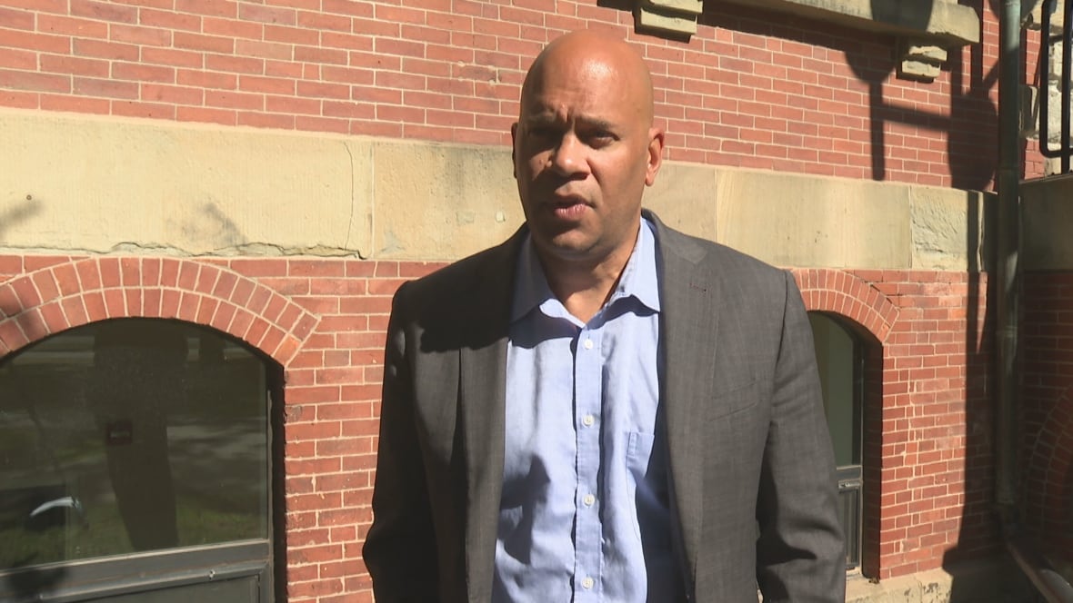 A bald man with a grey suit jacket over a pale blue button-down shirt stands in front of a brick building. 