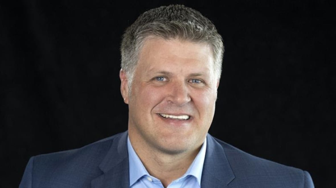 A smiling man wearing a blue collared shirt and blue blazer.