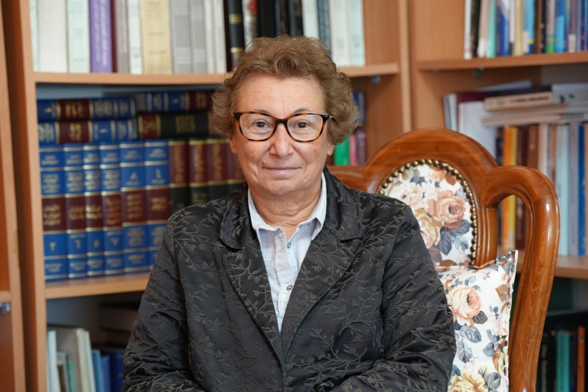 A woman in a black blazer sits on a chair. She's wearing brown glasses and has brown hair. Behind her are books on shelves.